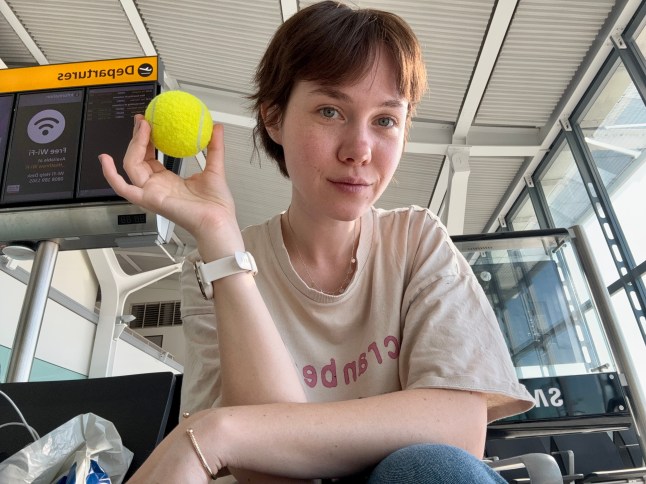 Maria Astardjieva holding a tennis ball in front of a departures gate at an airport.