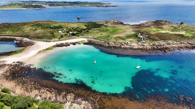 An aerial view of two sailboats at mooring at St Agnes in the Isles of Scilly. They are surrounded by turquoise water, and green and sandy coastline.