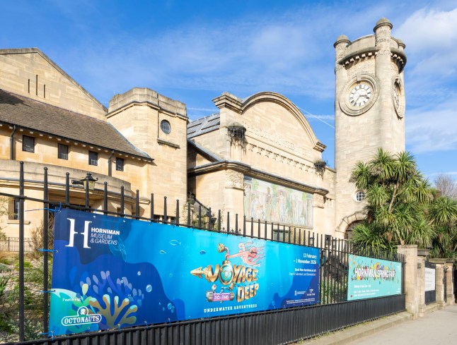 The exterior of the Horniman Museum in South London, with a clock tower, trees and a big poster on the front railings.