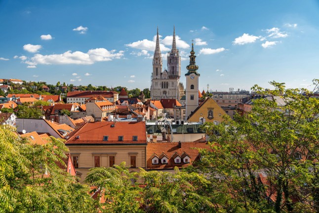 A cityscape of Zagreb, Croatia on a sunny day. There are green trees in the foreground, and brightly-coloured buildings, a cathedral and unusual clock tower behind them. The sky is light blue.