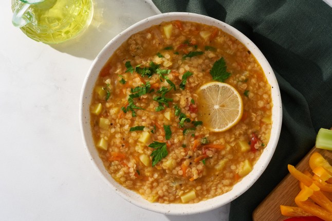 Bowl of hearty red lentil soup containing diced potatoes, carrots, and tomatoes. The dish is garnished with fresh chopped parsley and a slice of lemon, served in a speckled ceramic bowl on a white marble surface. A glass pitcher of oil and cut bell peppers are visible in the background.