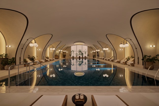 An indoor luxury swimming pool dimly lit with yellow lighting at Six Senses London. Lamps line the walls and sun loungers with luxurious white towels are dotted around the pool.