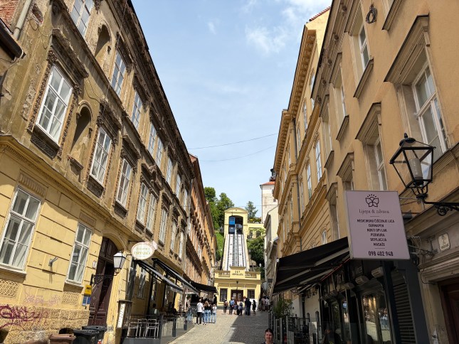 The funicular in Zagreb, Croatia, at the end of a narrow street with beige and stone-coloured shops and buildings.